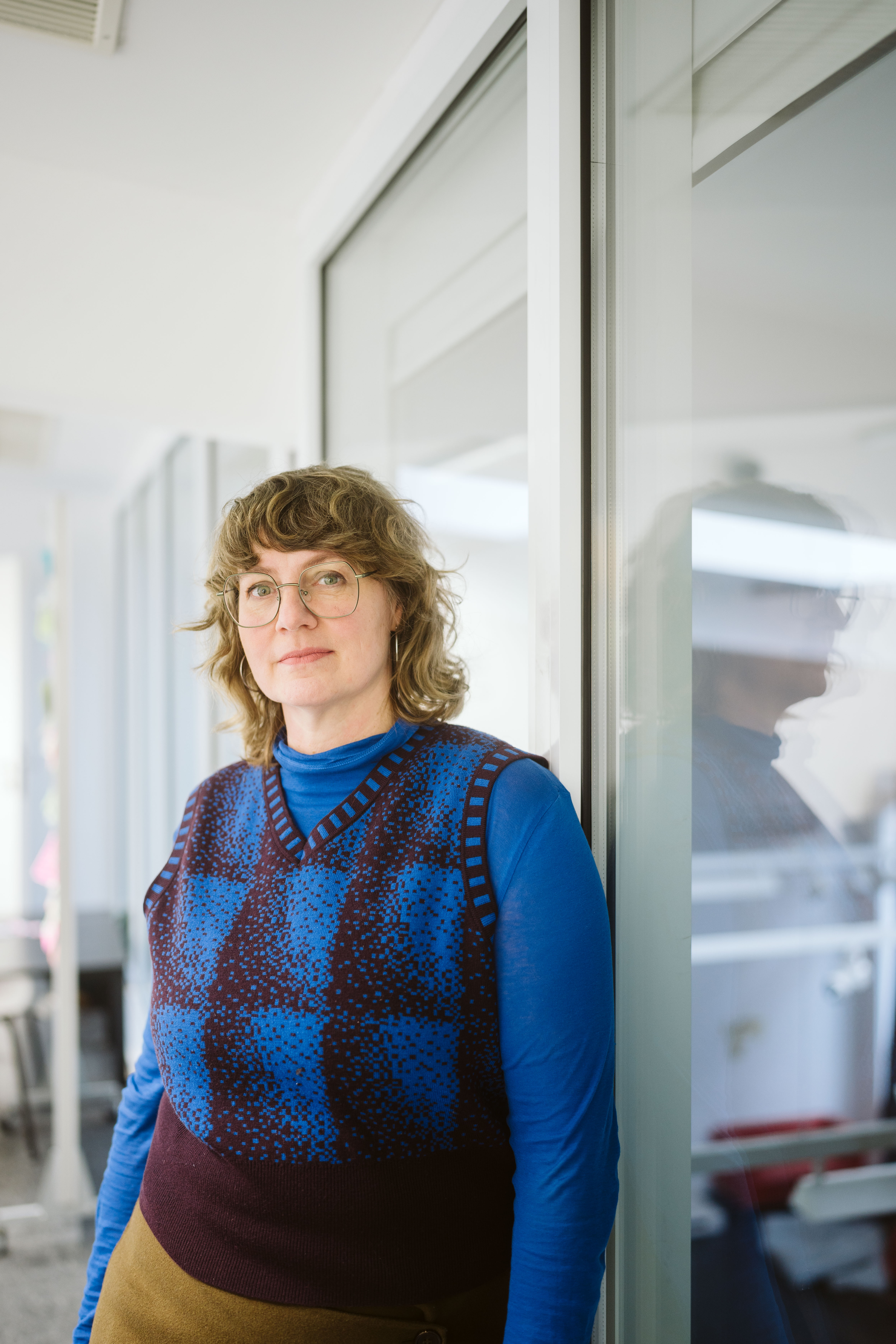 braun-blond haarige Frau in blauem Pullover und Cardigan die lehnend in die Kamera schaut.