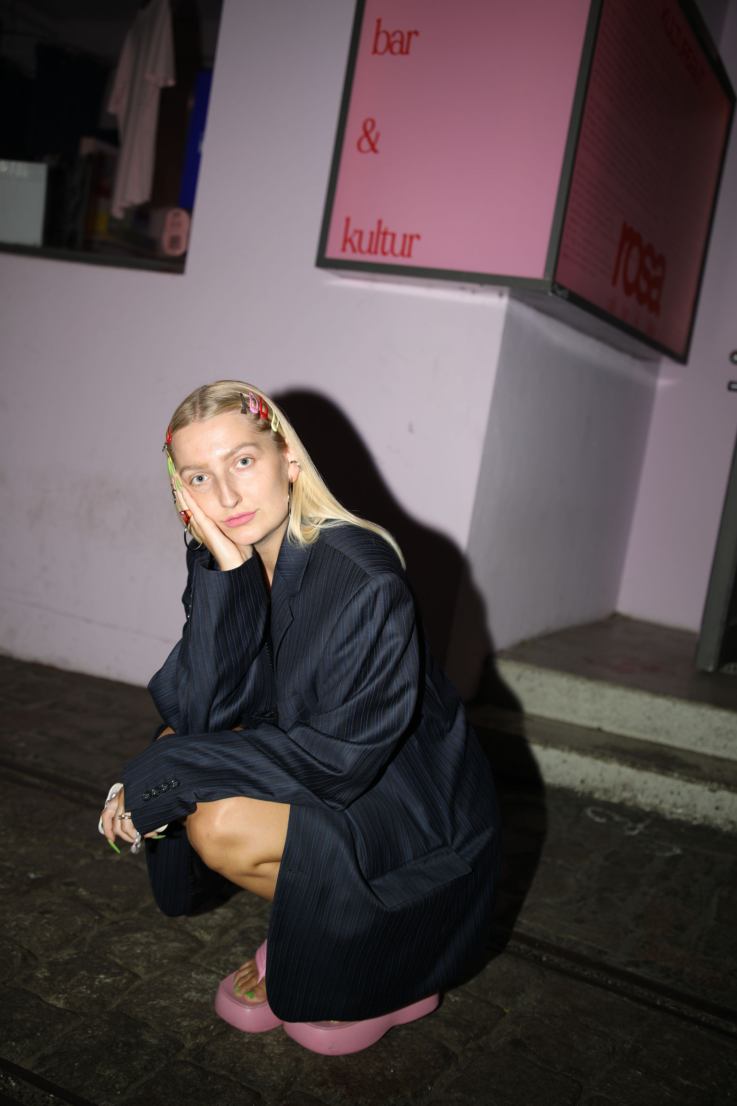 Junge Frau mit langen, blonden Haaren und bunten Haarspangen hockt auf dem Gehweg vor einem Gebäude mit der Aufschrift „bar & kultur“. Sie trägt ein übergroßes, dunkel gestreiftes Sakko und rosa Plateauschuhe. Ihr Gesicht liegt nachdenklich auf ihrer Hand. Die Szene ist bei Nacht aufgenommen und mit Blitz beleuchtet.