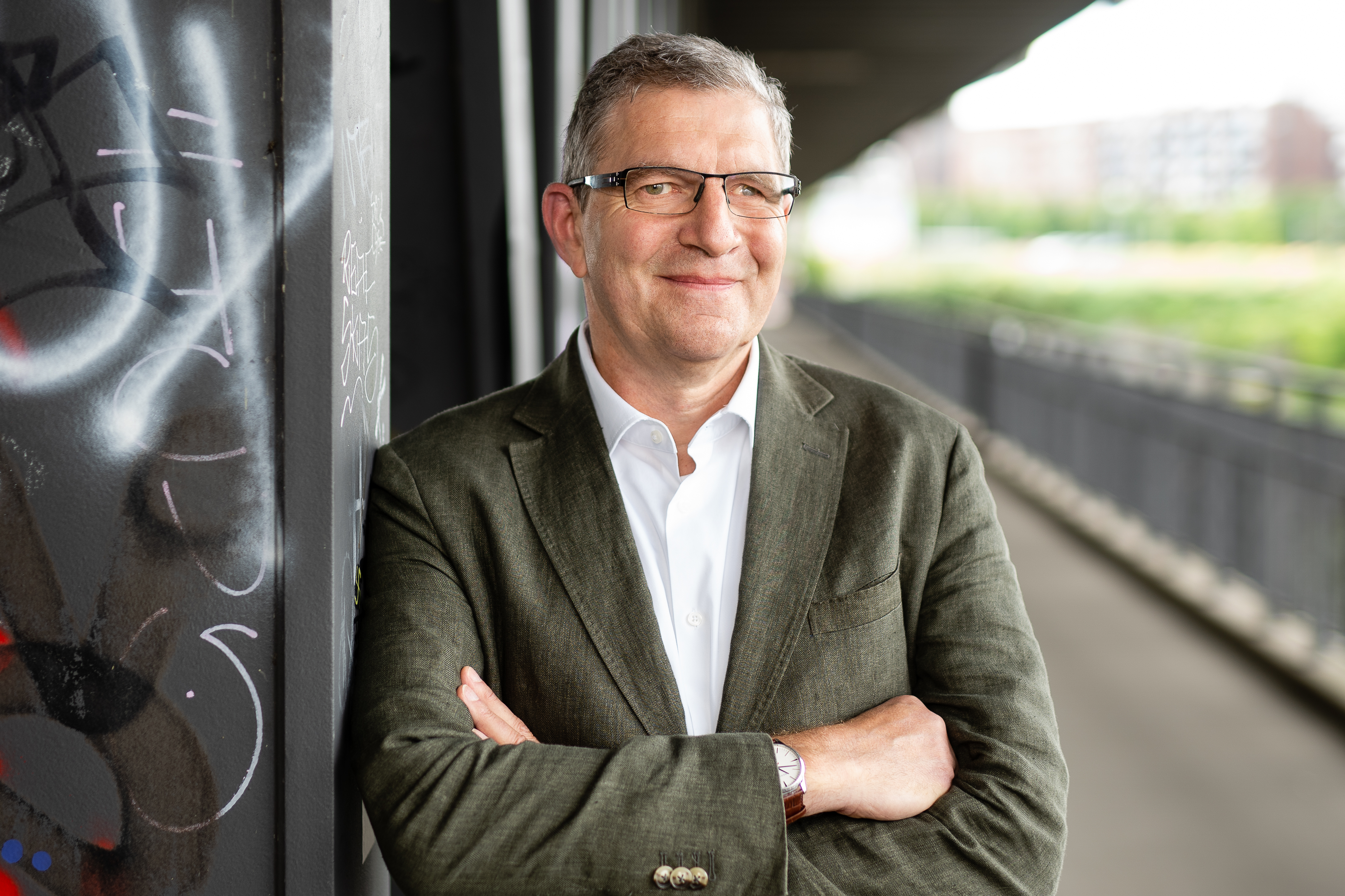 Älterer Mann mit kurzen grauen Haaren mit Grünem Sakko und weißem Hemd, lehnend an einer Wand mit einer Straße im Hintergrund.