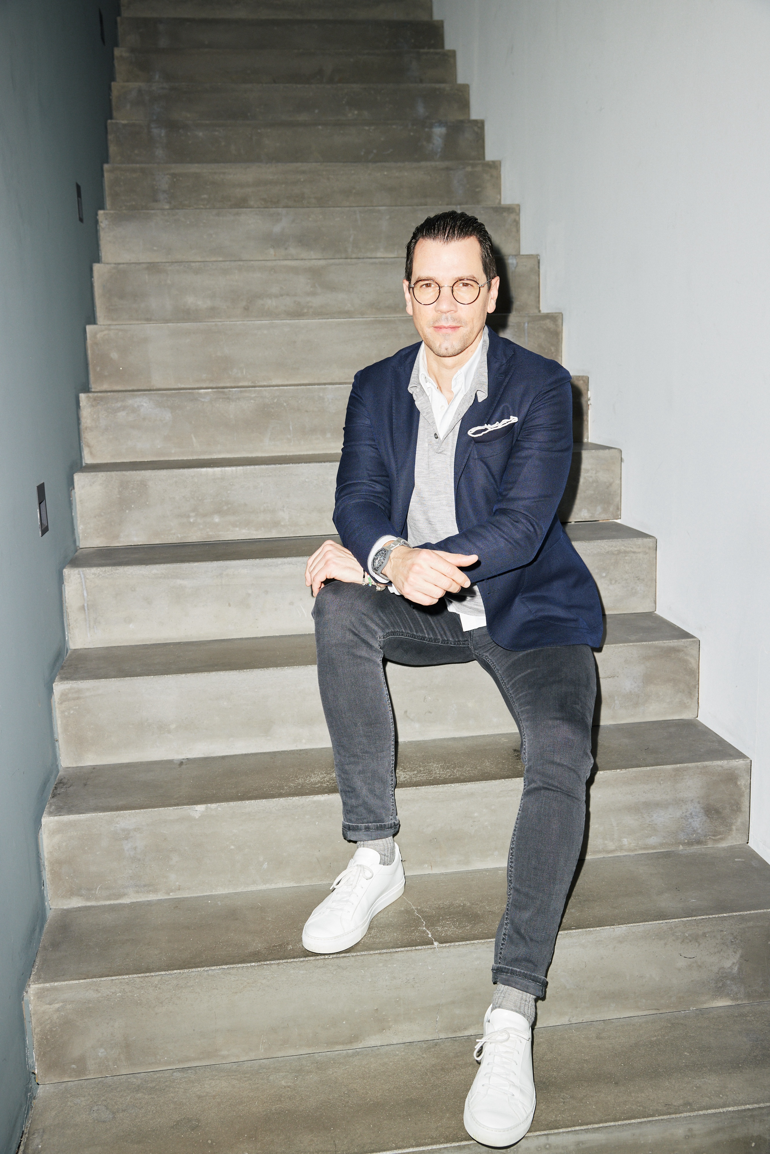 junger Mann wie er auf einer Betontreppe sitzt, beleuchtet von grellem Blitz in blauem Sakko und schwarzer Jeans wie er nett in die Kamera schaut