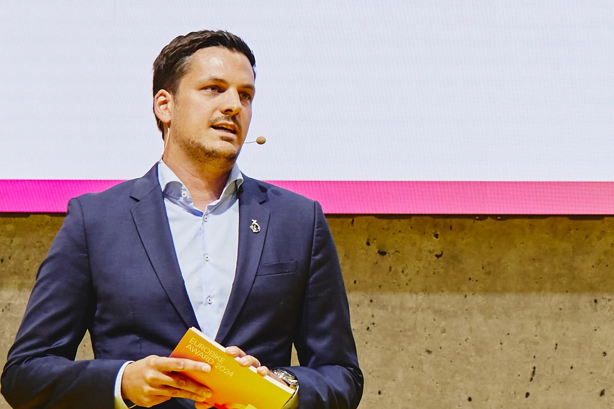 Moderator im dunkelblauen Anzug mit Headset-Mikrofon auf einer Bühne, hält Karten; Projektion mit pinker Leiste, Betonwand im Hintergrund.