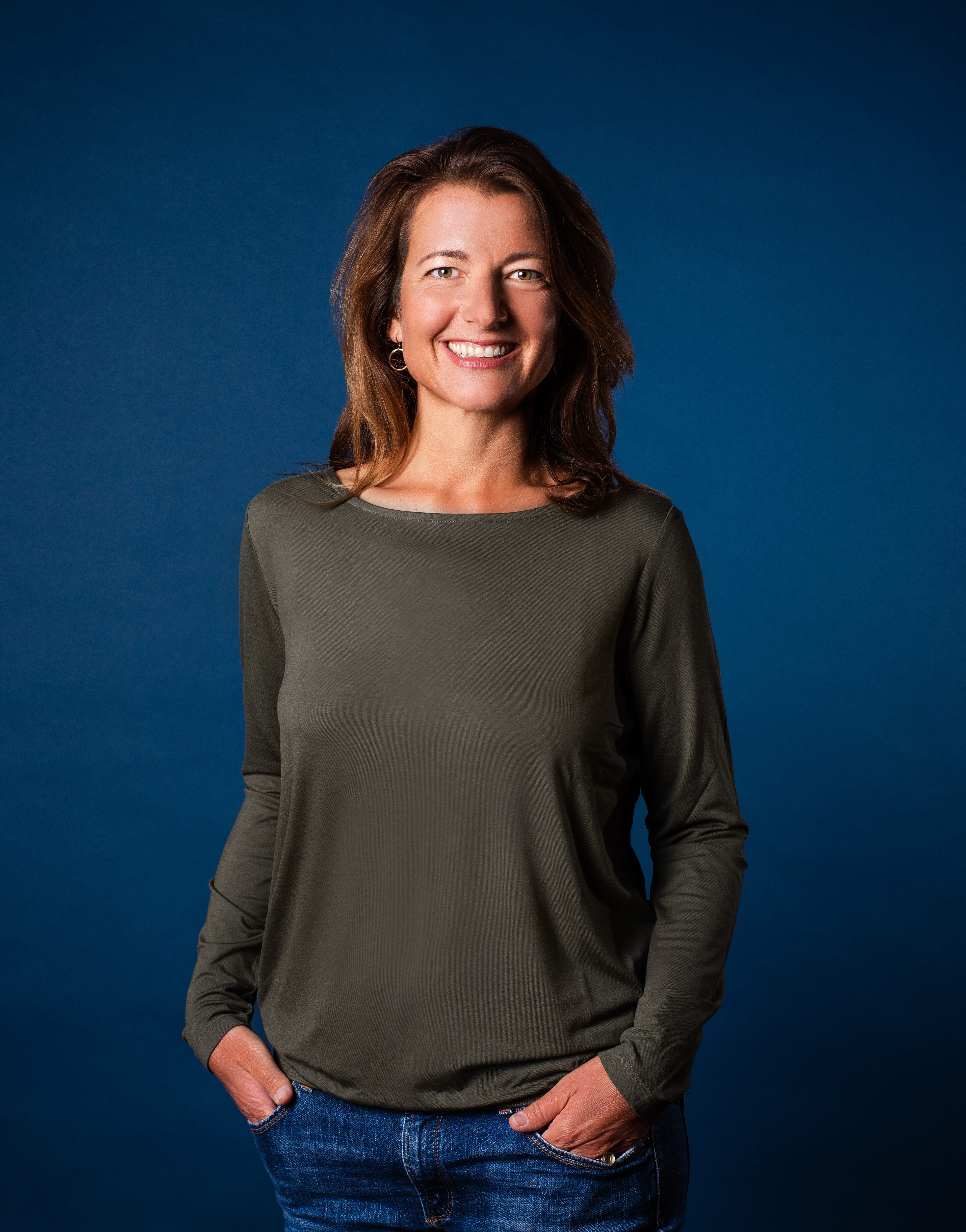 mittelalte Frau mit braunen Harren, grauem Shirt und blaue Jeans die vor einem dunkel blauen Studiohintergrund in die Kamera lächelt.