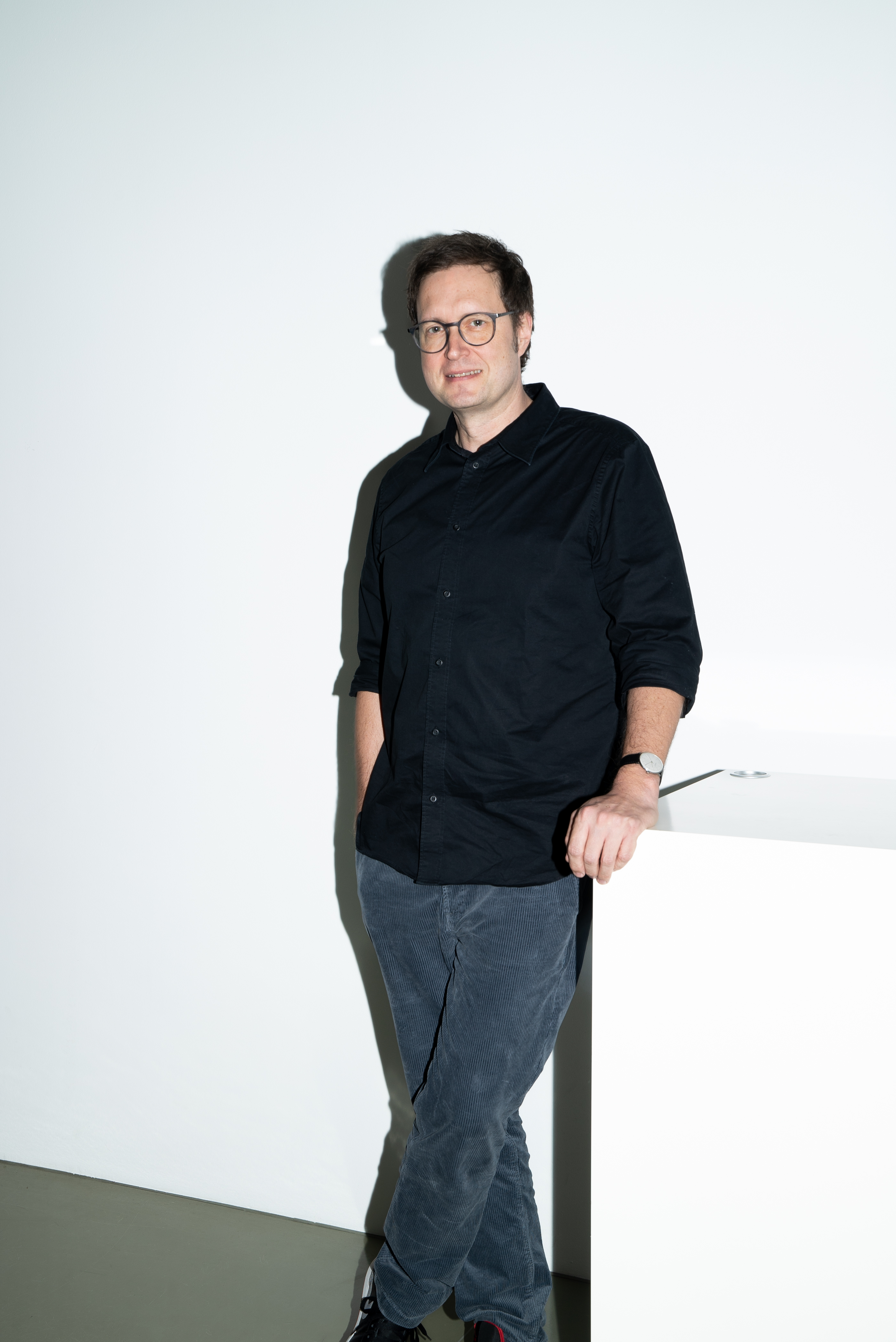Mittelalter Mann in schwarzem Pulli und blauer Hose, der vor einer weißen Wand steht und nett in die Kamera lächelt.