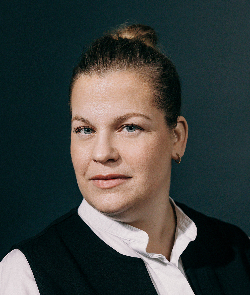 Malin Schuld ist eine Frau mit blonden Haaren, die zu einem strengen Dutt nach hinten gebunden sind. Sie hat einen breiten Mund und tief liegende, blaue Augen, mit denen sie direkt in die Kamera blickt. Sie trägt eine weiße Bluse und eine schwarze Veste darüber