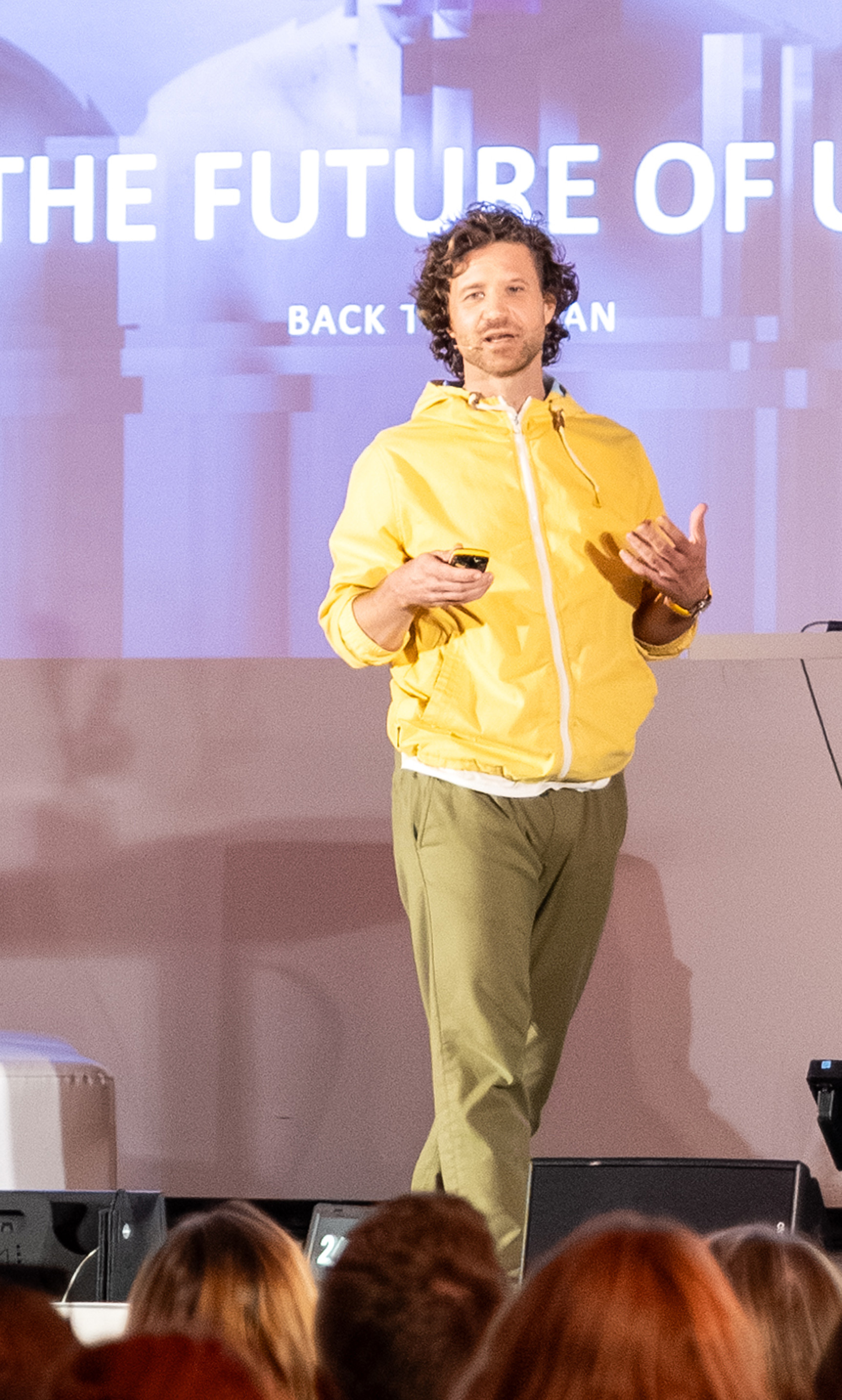 Braunhaariger Mann mit gelbem Hoodie und grüner Chino Hose beim vorsprechen vor einem Messepublikum in einer erklärenden und laufenden Bewegung festgehalten