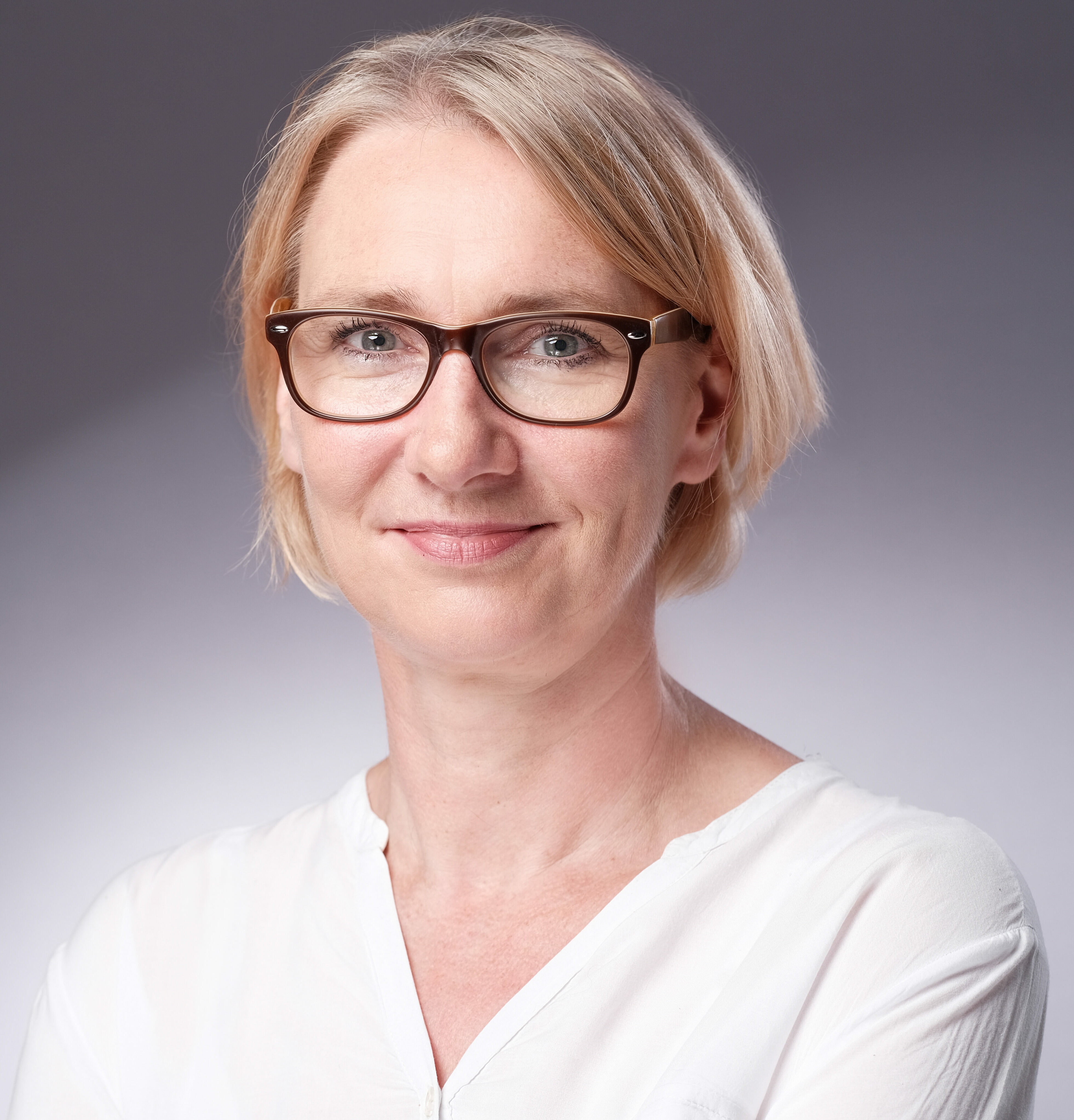 Porträt einer lächelnden Frau mit kurzem, blondem Haar und Brille. Sie trägt eine weiße Bluse und blickt direkt in die Kamera. Der Hintergrund ist neutral grau und leicht verlaufend ausgeleuchtet.
