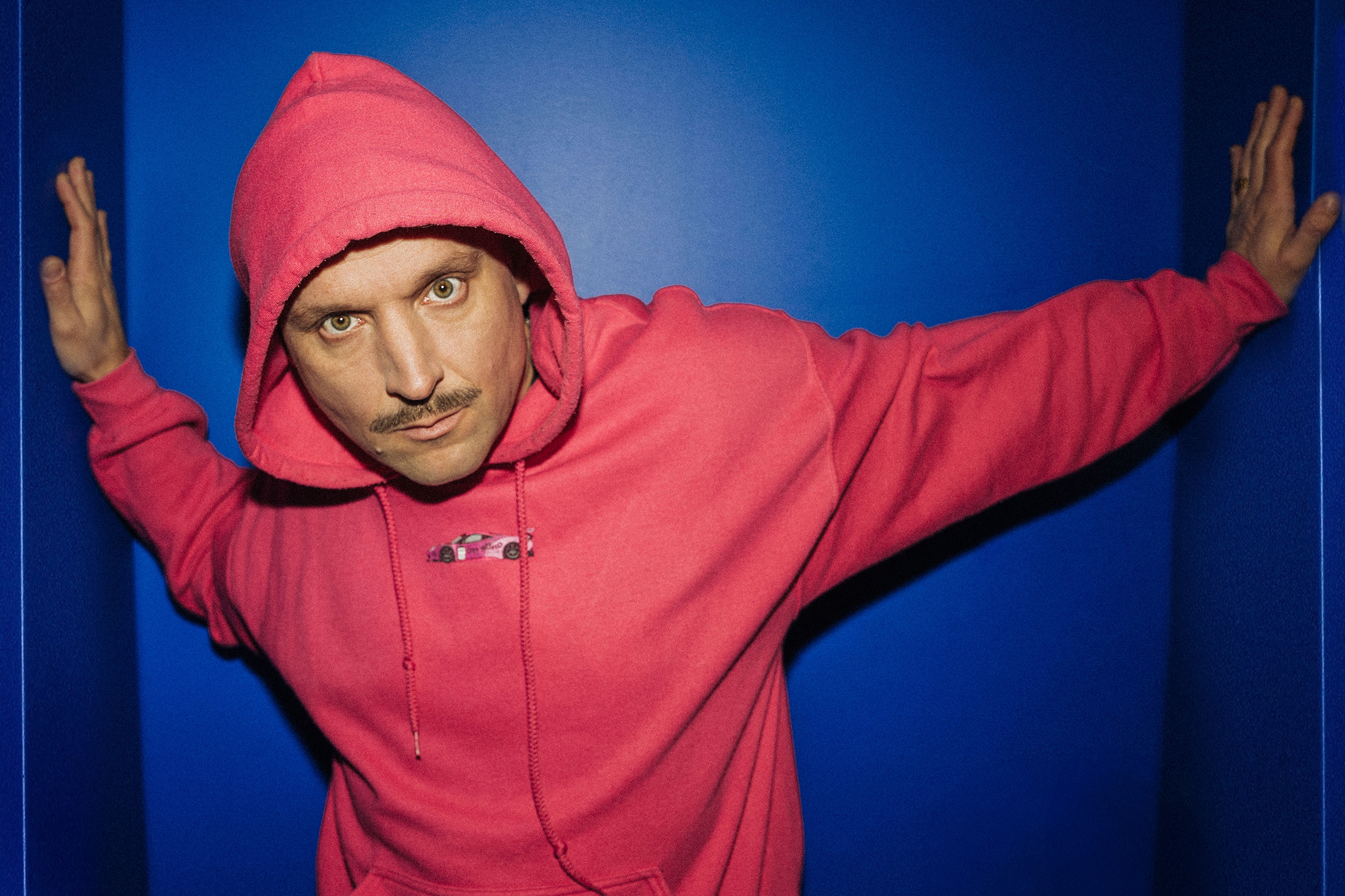 Ein weißer Mann mit Schnauzer welcher grimmig in die Kamera schaut. Er trägt einen pinken Pulli vor einem blauen Hintergrund.